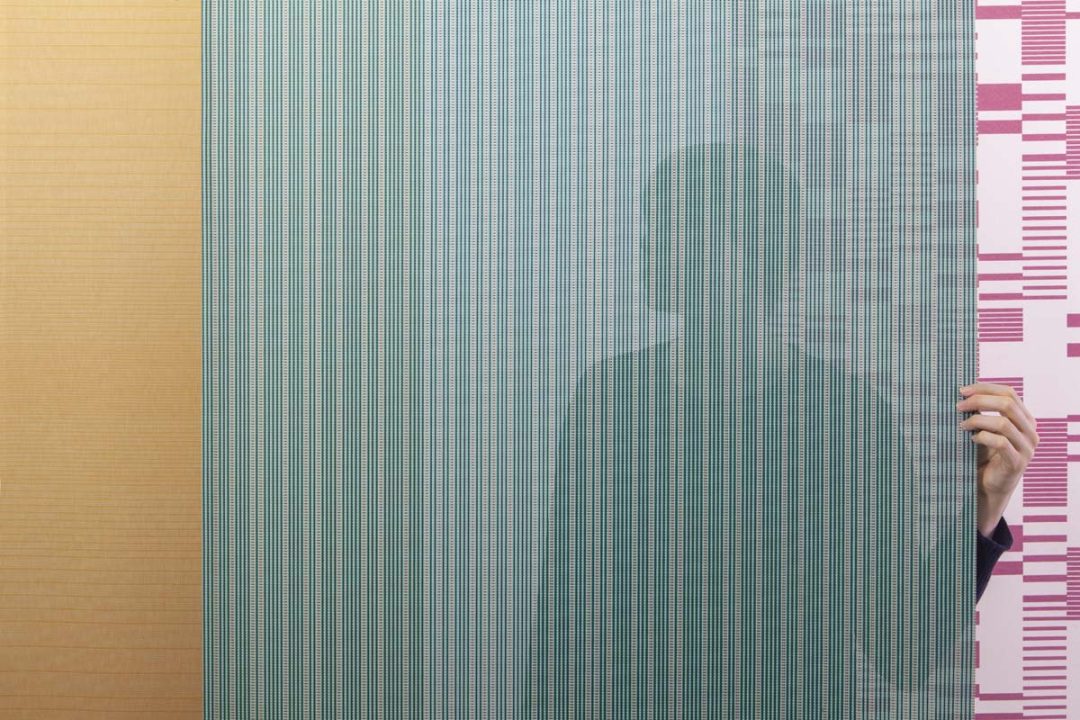 El estudio RAW COLOR ha diseñado tres modelos de telas para cortinas vertisolfabrics: Hatching, Density and Rows. La imagen muestra la transparencia del tejido con una persona detrás.